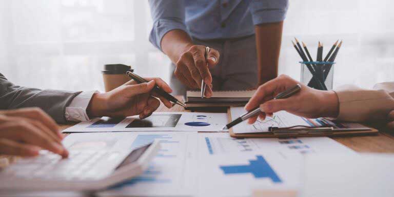 3 hands with pens reviewing spreadsheet data