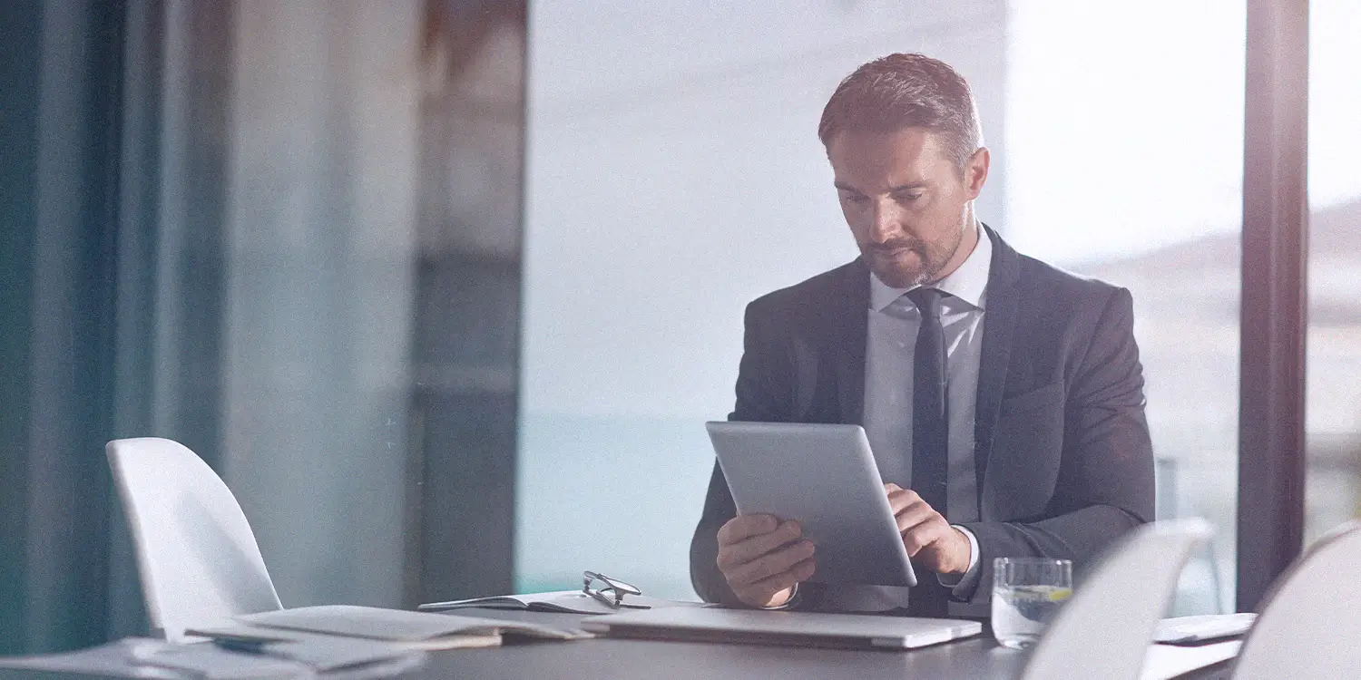 A man in a suit and tie looks at a tablet while sitting at a conference table in an office
