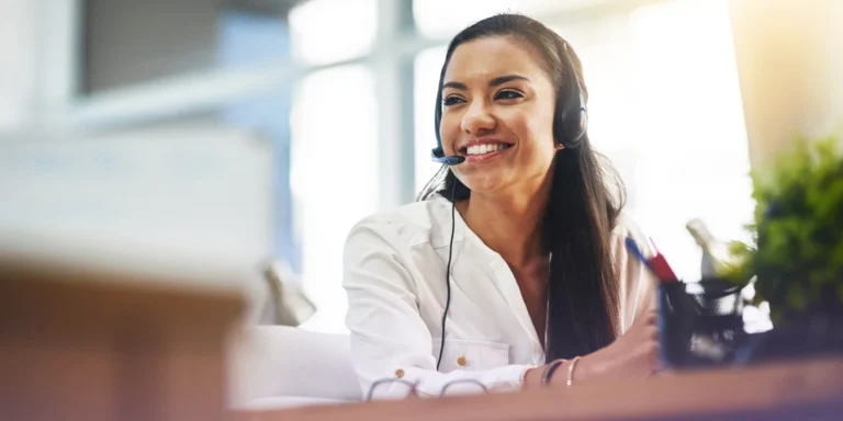 A smiling female contact center agent wears a headset and takes a call from a customer