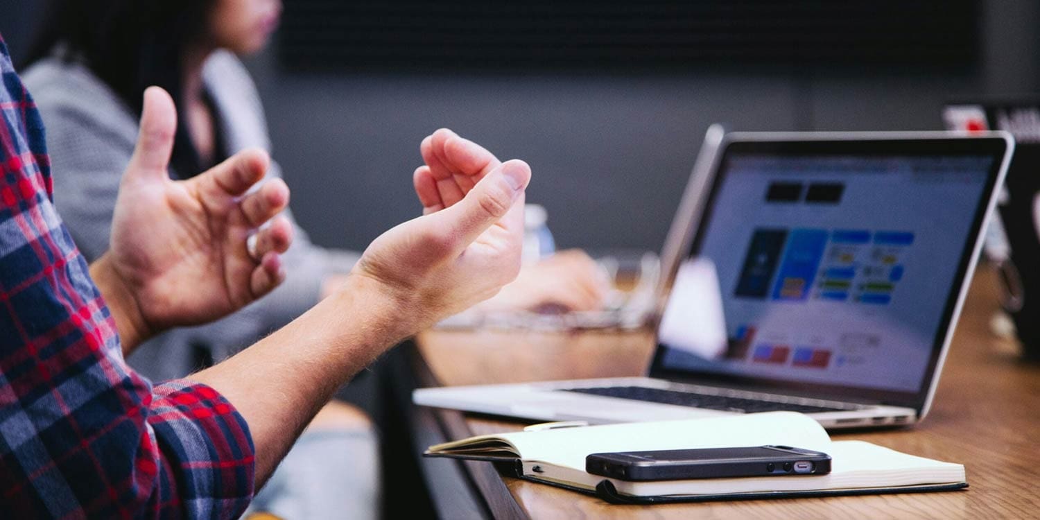 Personne gesticulant lors d'une réunion, avec un ordinateur portable affichant des maquettes de design, un smartphone et un carnet sur la table, et un collègue flou en arrière-plan.