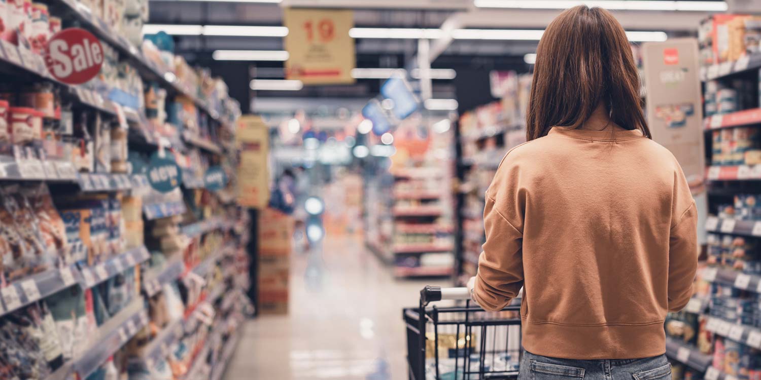 A woman pushing a shopping cart through the aisles of a supermarket, surrounded by shelves stocked with groceries.