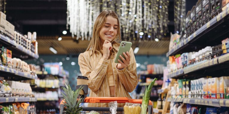 A woman in the aisle of a store with a shopping cart in front of her, looks at her smartphone