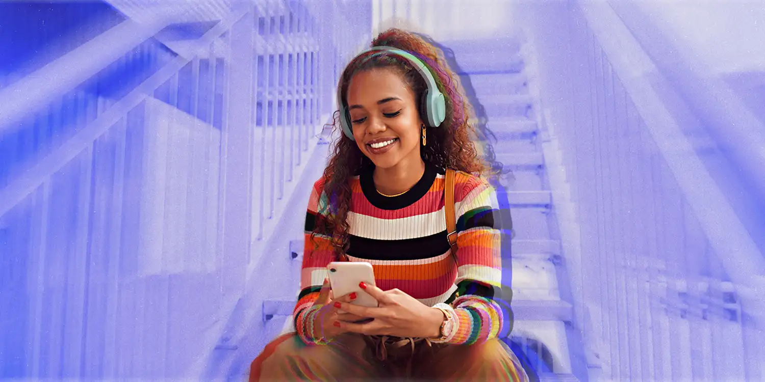 A female in her 20s smiles while sitting on stairs and wearing headphones and looking at her smartphone