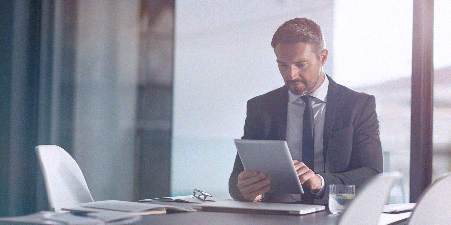 A man in a suit and tie looks at a tablet while sitting at a conference table in an office