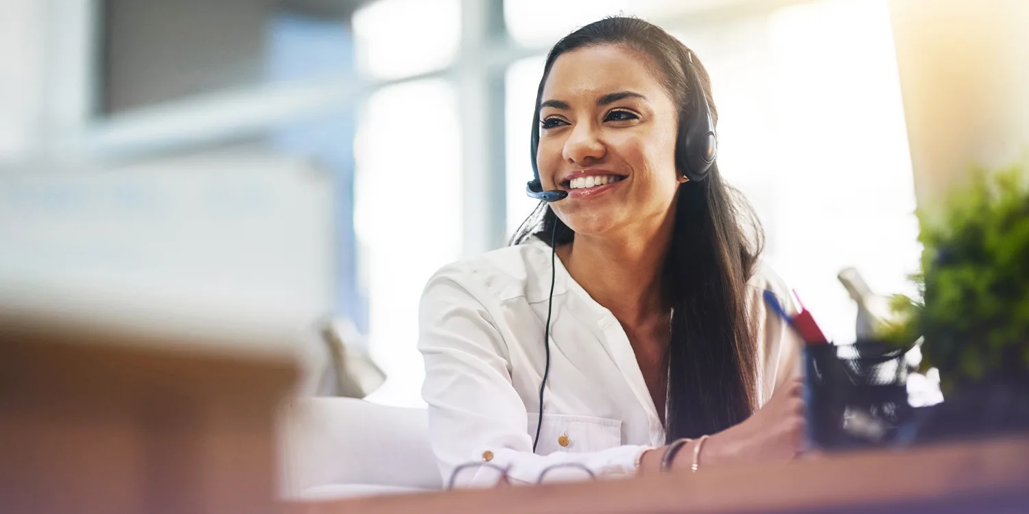 A smiling female contact center agent wears a headset and takes a call from a customer