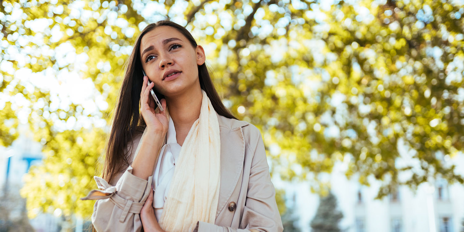 A female holds a mobile phone to her ear while standing outside under a tree