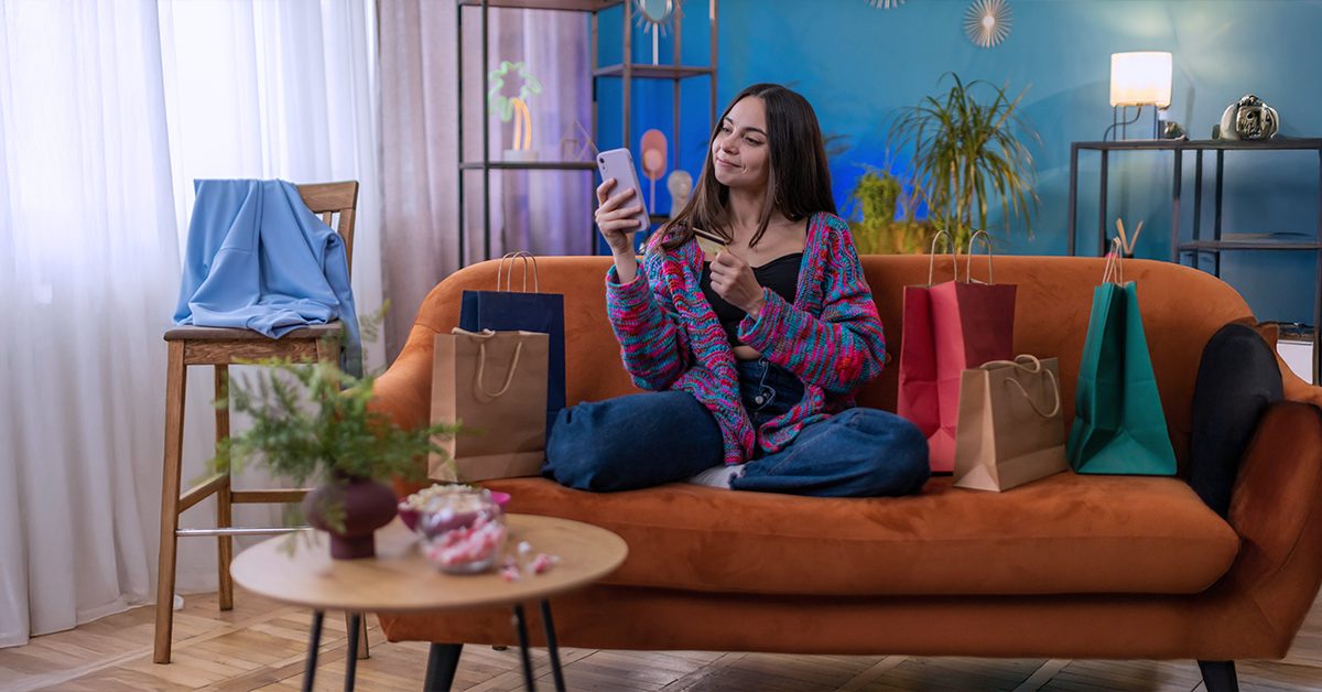 A female sits on a sofa in her home while shopping online on her smartphone and gift bags sitting on the right side of the sofa