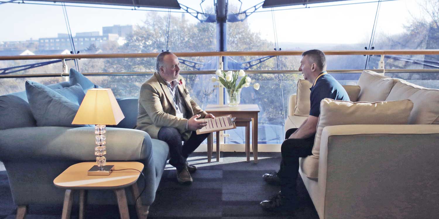 A supervisor coaches an employee while sitting opposite on two sofas in an office area
