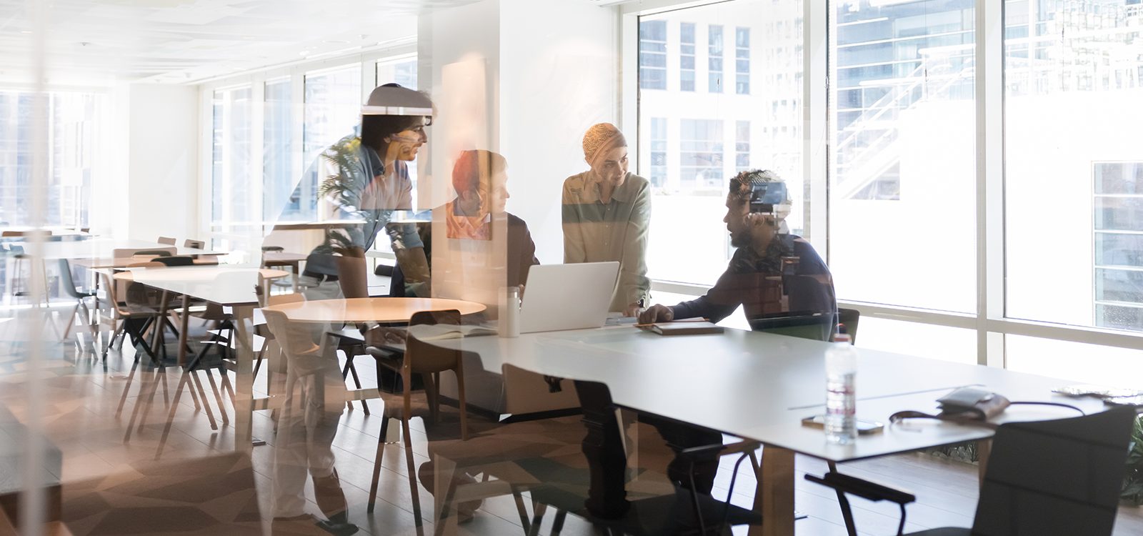 Four people meeting in an office conference room are gathered around a laptop and look like they are planning