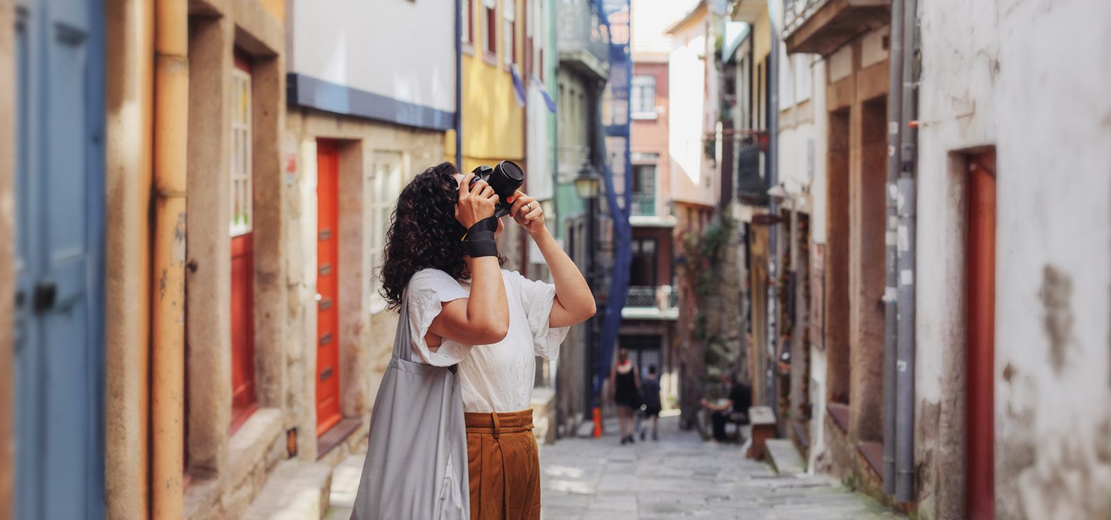 A woman takes a photo with a high-end camera while standing in a walkway between buildings with colorful doorways