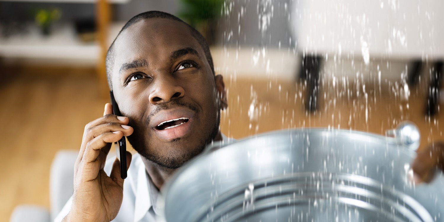 Closeup of a man speaking to someone on his smartphone while holding a bucket to catch water leaking from a ceiling