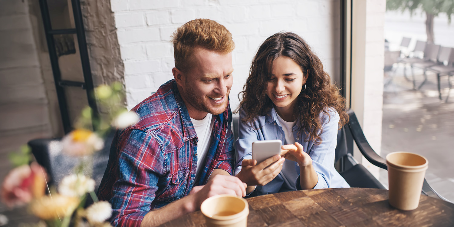 A man and woman sit at a dining table and look at the phone the lady is holding