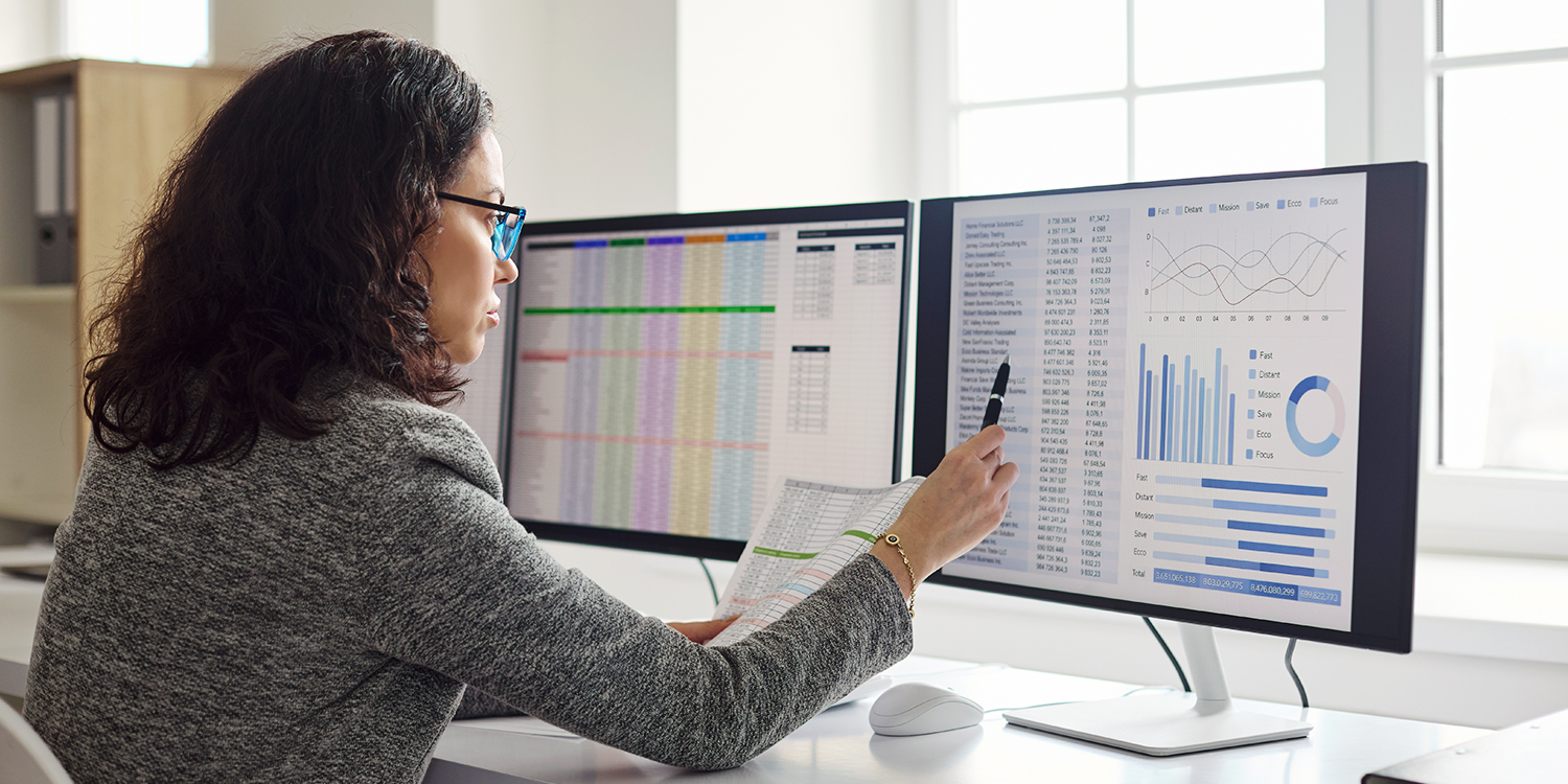 A woman sitting at a desk looks at data on two computer monitors